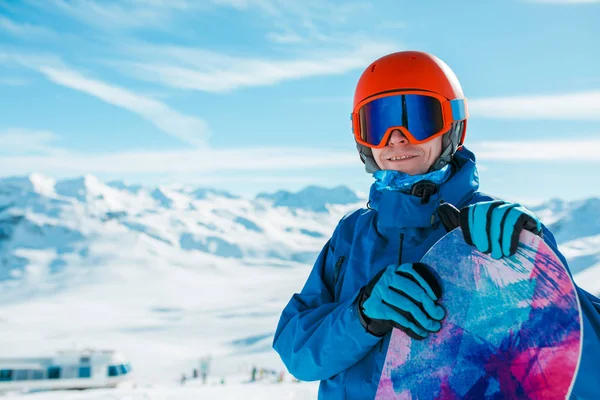 Obrázek sportive člověka v helmě, při pohledu na fotoaparát s snowboard na pozadí zasněženého kopce — Stock fotografie