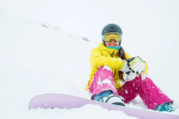 Foto de mujer deportiva con casco y máscara con snowboard sentado en la pendiente de la colina nevada —  Fotos de Stock