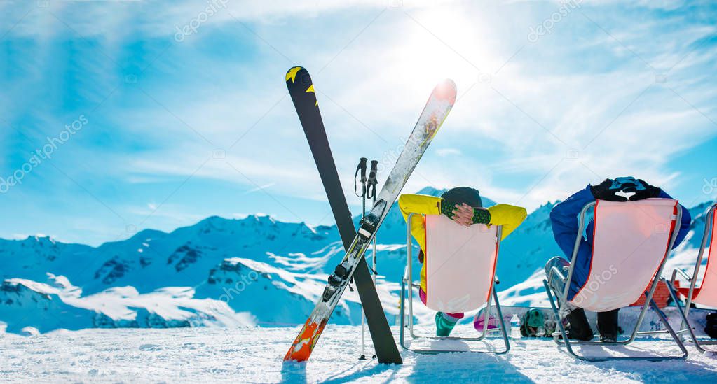 Image from back of vacationers in armchair, skis, sticks in snowy resort