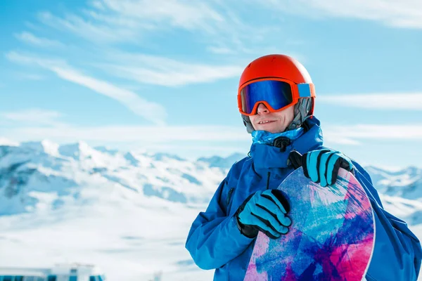 Foto de hombre deportivo en casco mirando a la cámara con snowboard en el fondo de la colina nevada —  Fotos de Stock
