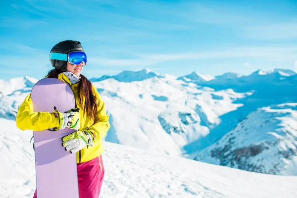 Foto de morena sonriente en casco y máscara con snowboard sobre fondo de colinas nevadas — Foto de Stock