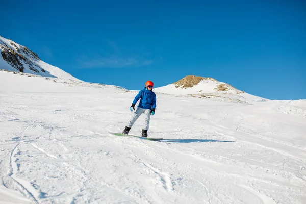 スノーボードの雪が多い斜面に乗ってヘルメットの青いジャケットを着てスポーツ男のイメージ — ストック写真