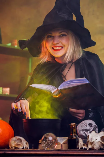 Imagem de bruxa sorrindo loira em chapéu preto com poção de fabricação de livros em panela — Fotografia de Stock