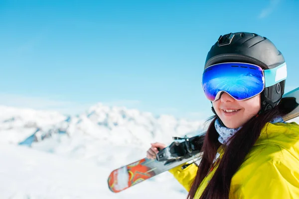 Portrait of sporty woman in helmet with skis on her shoulder against background of winter hills — Stock Photo, Image