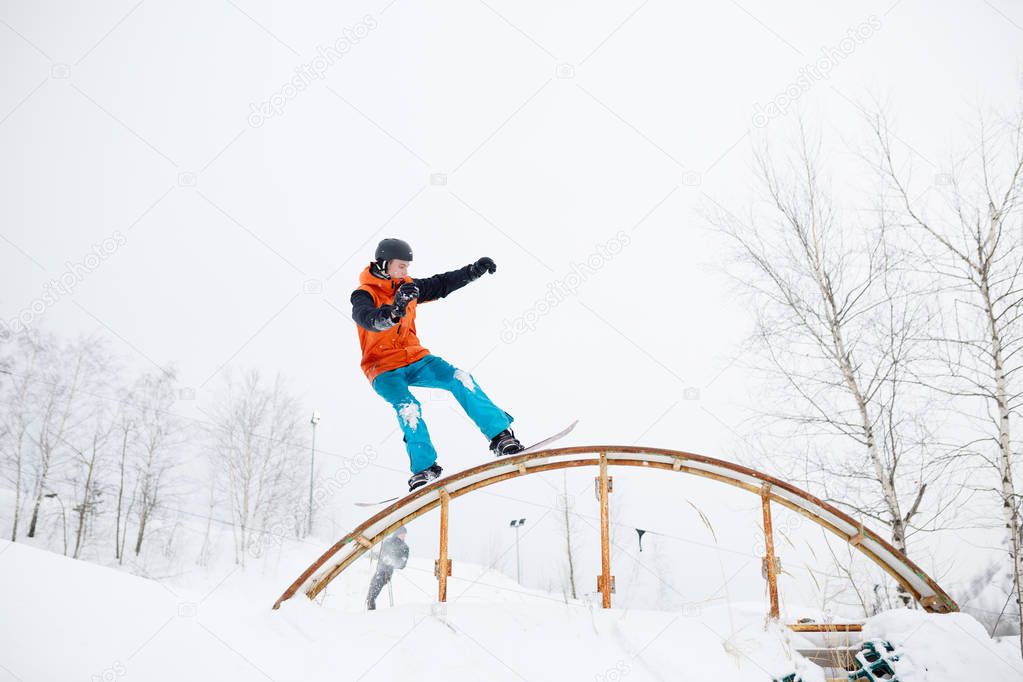 Photo of young sportive man skiing on snowboard with springboard against background of trees