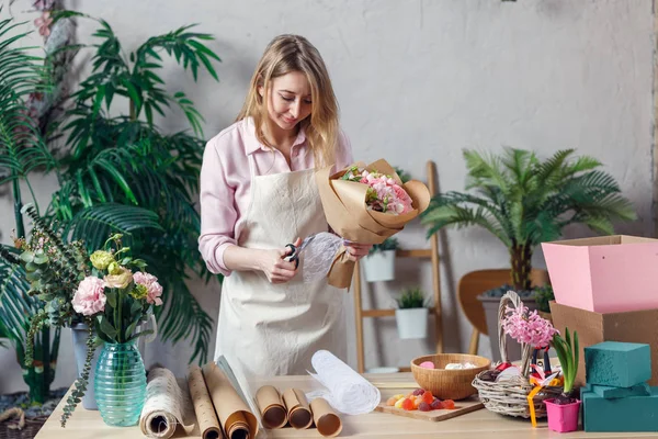 Foto de florista rubia en delantal con ramo con papel kraft en la mesa con papel, mermelada — Foto de Stock