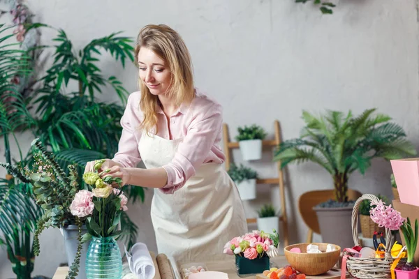 Фото флористки в фартуке с цветами в вазе за столом с зефиром, мармеладом — стоковое фото