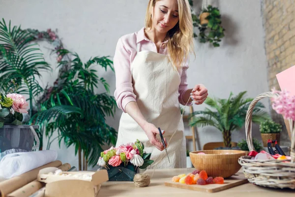 Imagen de mujer florista con cuerda para cortar tijeras en la mesa con ramo, mermelada, malvavisco — Foto de Stock