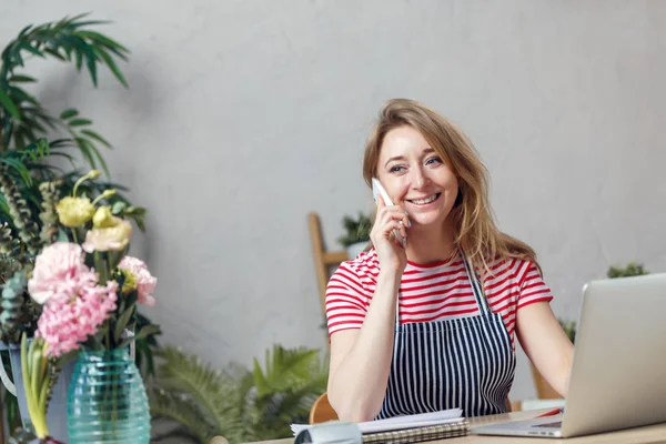 Foto de la florista hablando por teléfono mientras está sentada en la mesa con el ordenador — Foto de Stock