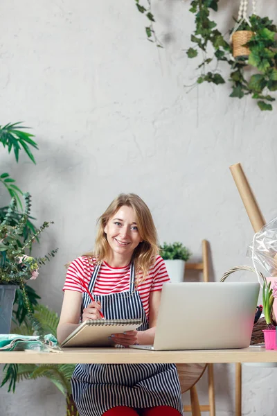 Foto de mujer floral sonriente con bloc de notas en la mesa con el ordenador portátil — Foto de Stock