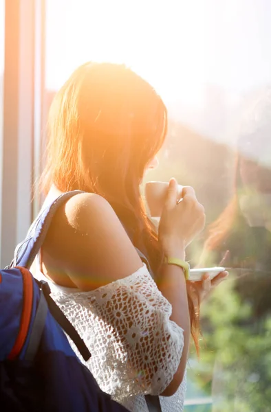 Image of long-haired pensive woman with cup of tea and backpack by window — Stock Photo, Image