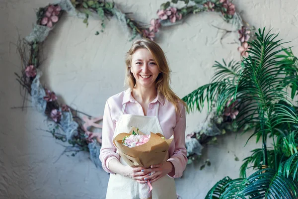 Foto de florista sonriente con ramo en las manos sobre el fondo del paisaje de flores en forma de corazón — Foto de Stock