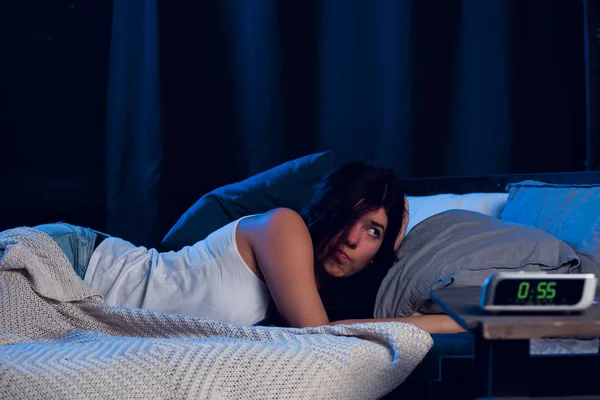 Foto, joven morena con insomnio tumbada en la cama al lado del reloj —  Fotos de Stock