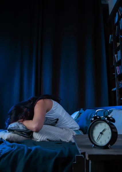 Фото незадоволеної брюнетки з безсонням сидить на ліжку поруч з будильником — стокове фото