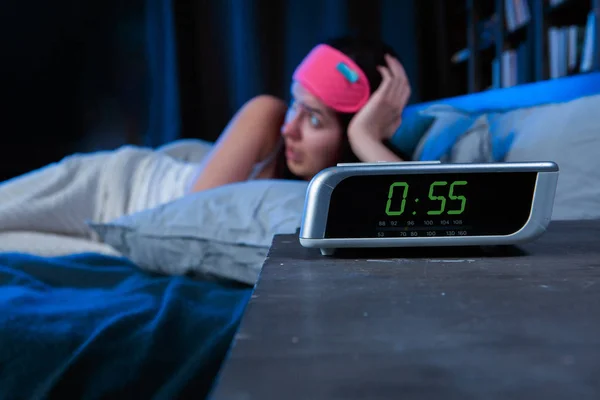 Изображение женщины с бессонницей с розовой повязкой для глаз, лежащей на кровати рядом с часами — стоковое фото