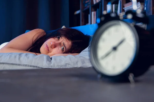 Foto de morena con insomnio tumbada en la cama junto al despertador por la noche —  Fotos de Stock