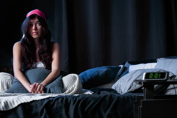 Photo of young brunette with insomnia with pink bandage for eyes sitting on bed next to clock — Stock Photo, Image