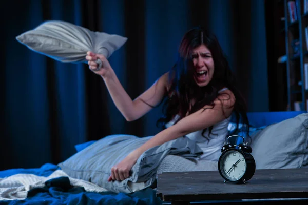 Imagen de mujer insatisfecha con insomnio arroja almohada sentada en la cama junto al despertador —  Fotos de Stock