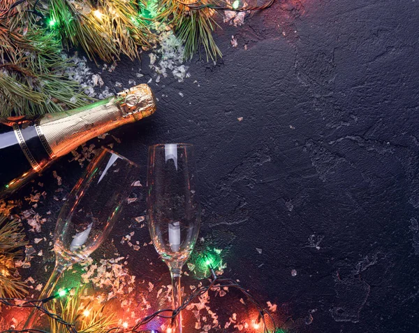 Picture of branches of fir, , burning garlands, champagne bottles, two wine glass on black background — Stock Photo, Image