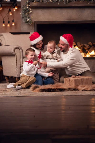 Imagen de los padres en gorra de Santas con dos hijos sentados en el piso junto a la chimenea en la habitación —  Fotos de Stock