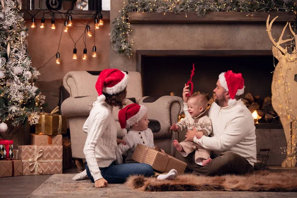 Imagen de los padres en gorra de Santas con dos hijos sentados en el suelo junto a la chimenea junto a un ciervo con guirnalda en la habitación —  Fotos de Stock