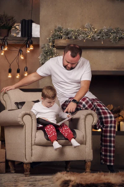 Foto de padre con hijo con libro sentado en sillón en la habitación con el interior de Navidad . —  Fotos de Stock