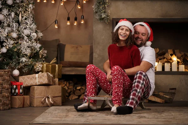 Imagen de pareja joven en gorras de Santas sentada en trineo en la habitación — Foto de Stock