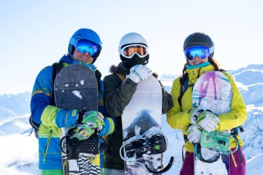 Kar tesisinde ellerinde kask ve snowboard giyen üç sporcunun fotoğrafı..