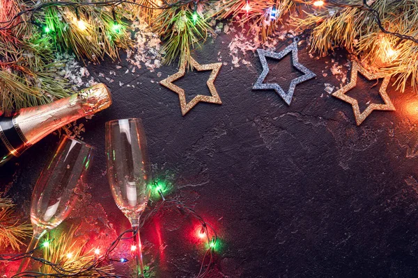 Photo of branches of fir, Christmas decorations blue and gold toys, burning garlands, champagne bottles, two wine glass on black background — Stock Photo, Image