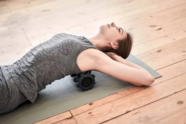 Brunette doing yoga lying on massager on mat on wooden floor in gym — Stock Photo, Image