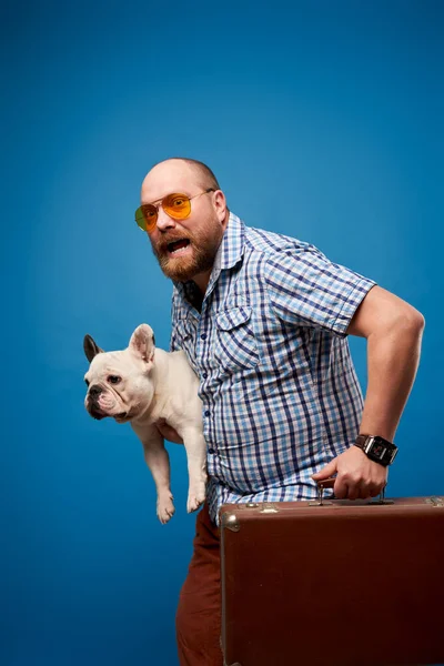 Szakállas férfi szemüvegben kutya és poggyász üres kék háttér — Stock Fotó