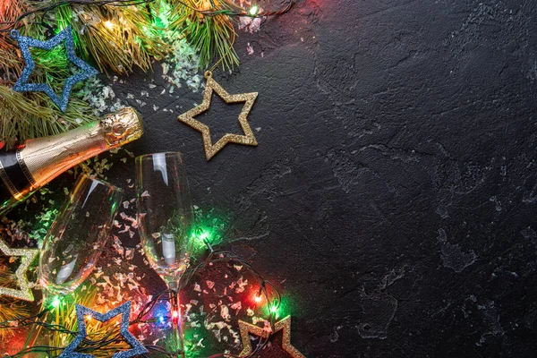 Imagen de ramas de abeto, adornos navideños juguetes azules y dorados, guirnaldas ardientes, botellas de champán, dos copas de vino sobre fondo negro — Foto de Stock