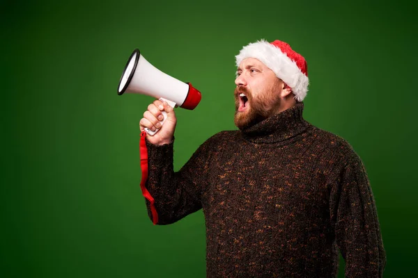 Mann mit Weihnachtsmütze und Pullover schreit in Ochsenhorn auf leerem grünen Hintergrund — Stockfoto