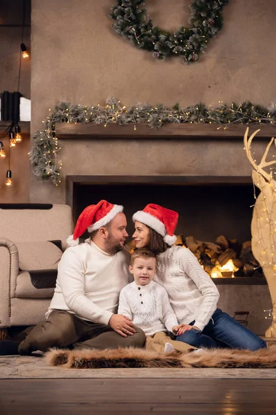 Foto de hombre y mujer en gorra de Santas con hijo sentado en el suelo sobre el fondo de la chimenea —  Fotos de Stock