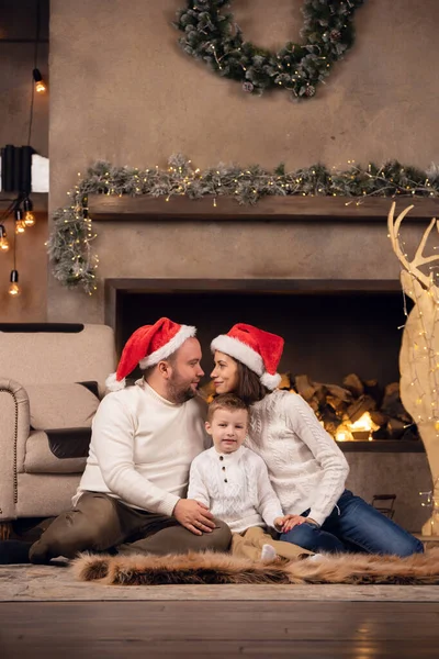 Imagem de homem e mulher em boné Santas com filho sentado no chão no fundo da lareira — Fotografia de Stock