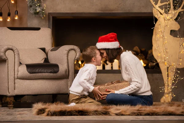 Foto van brunette vrouw in Santas cap met zoon zittend op de vloer op de achtergrond van open haard — Stockfoto