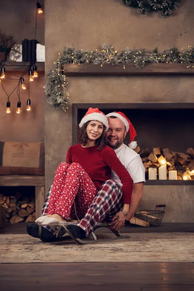 Photo de couple heureux dans les casquettes Santas assis sur traîneau dans la chambre — Photo