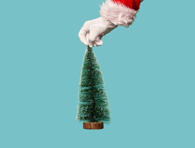 Santas hand in white gloves holding artificial Christmas tree on empty blue background. clipart