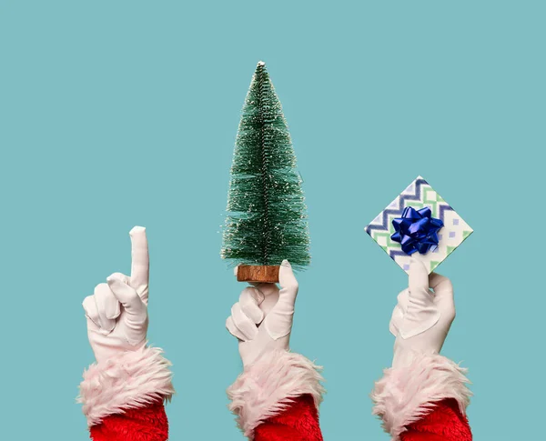 Три руки Санты в белых перчатках с искусственной елкой, подарок на пустом синем фоне . — стоковое фото