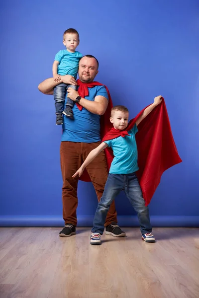 Papá y dos hijos superhéroes en capas rojas sobre fondo azul vacío — Foto de Stock