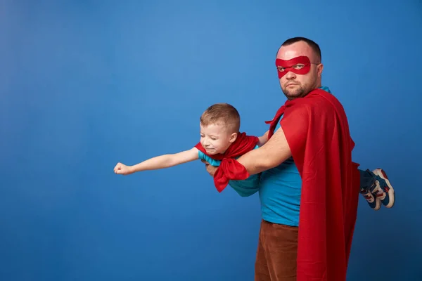 Papa in Maske und fliegender Sohn Superhelden in roten Mänteln auf blauem Hintergrund — Stockfoto