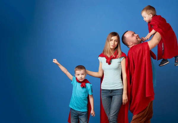 Szczęśliwa mama i tata, dzieci superbohaterowie w czerwonych płaszczach na czystym niebieskim tle — Zdjęcie stockowe
