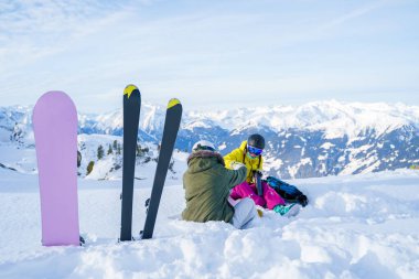 Kayak resimleri, kışın kayak merkezinde oturan termoslu iki kadın sporcunun arka planına karşı snowboard.