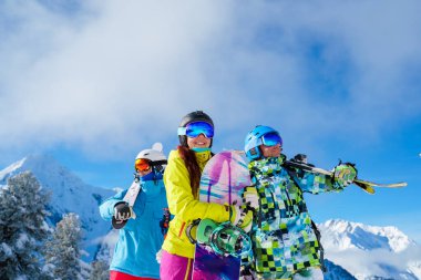 Snowboard ve kayak yapan erkek ve kadınlar farklı yönlere bakıyor. Dağların ve bulutlu gökyüzünün arka planında kar üzerinde duruyorlar..
