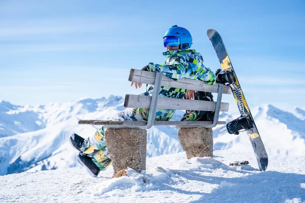 Photo from back of sportsman sitting on bench next to snowboard in ski resort — ストック写真