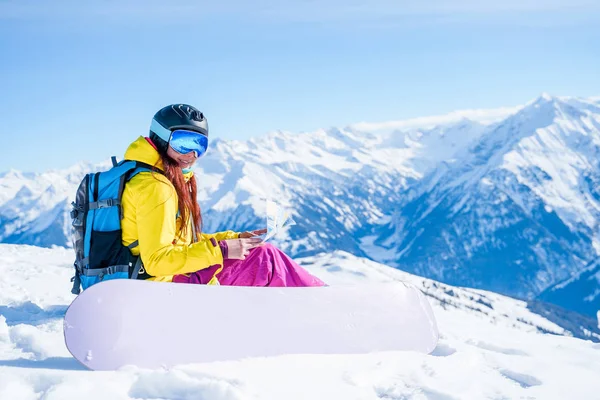 Foto de niña snowboarder en casco con mapa en sus manos sentadas en la ladera de la montaña —  Fotos de Stock