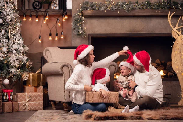 Imagem de pais felizes em boné Santas com dois filhos sentados no chão pela lareira no quarto — Fotografia de Stock