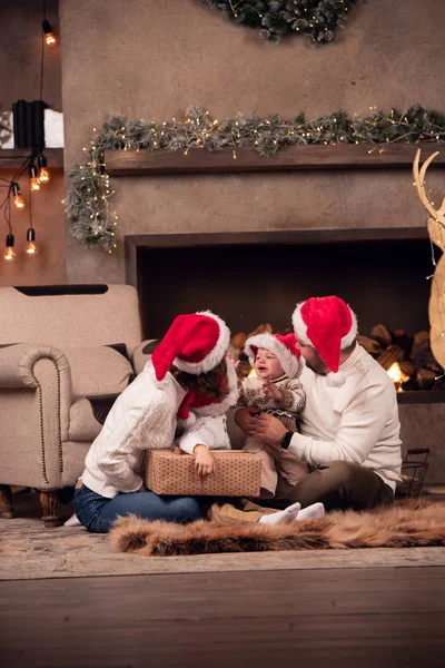 Foto de padres felices en gorra de Santas con dos hijos sentados en el piso junto a la chimenea en la habitación —  Fotos de Stock