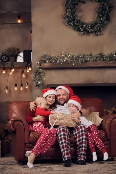 Imagen de padres jóvenes en gorras Santas e hijos en sofá de cuero rojo en estudio — Foto de Stock