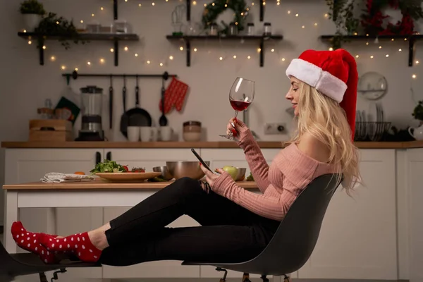 Şarap kadehi ve telefonla Noel Baba şapkalı bir kız mutfakta yanan çelenklerle oturuyor. Yeni yıl kavramı — Stok fotoğraf